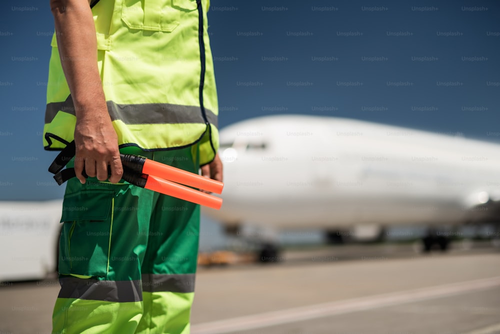 Work outfit. Close up of male arm holding sticks for signaling. Passenger plane and runway on blurred background
