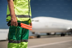 Work outfit. Close up of male arm holding sticks for signaling. Passenger plane and runway on blurred background