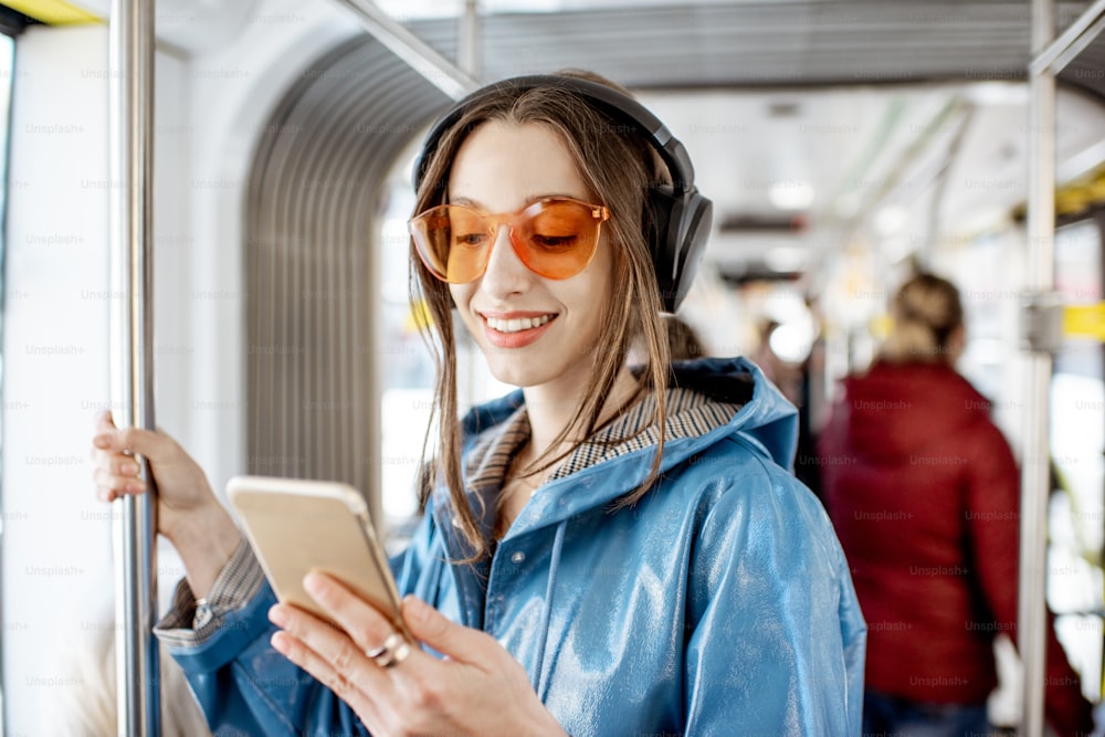 Junge stilvolle Frau, die öffentliche Verkehrsmittel benutzt, mit Kopfhörern und Smartphone steht, während sie sich in der modernen Straßenbahn bewegt