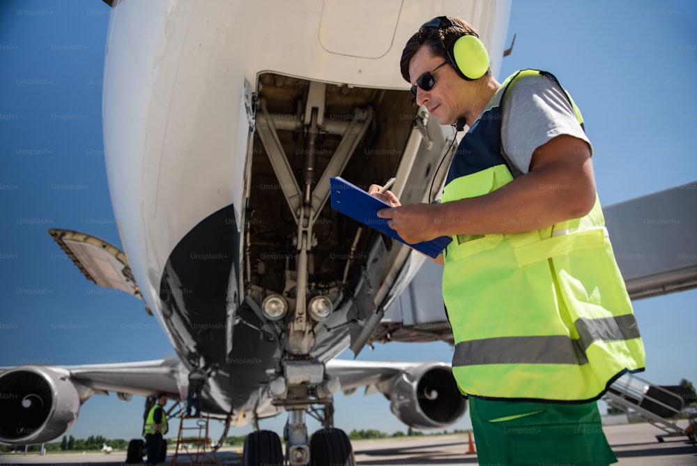 Doing paperwork. Low angle portrait of man in sunglasses writing on clipboard while standing near commercial jet with open door