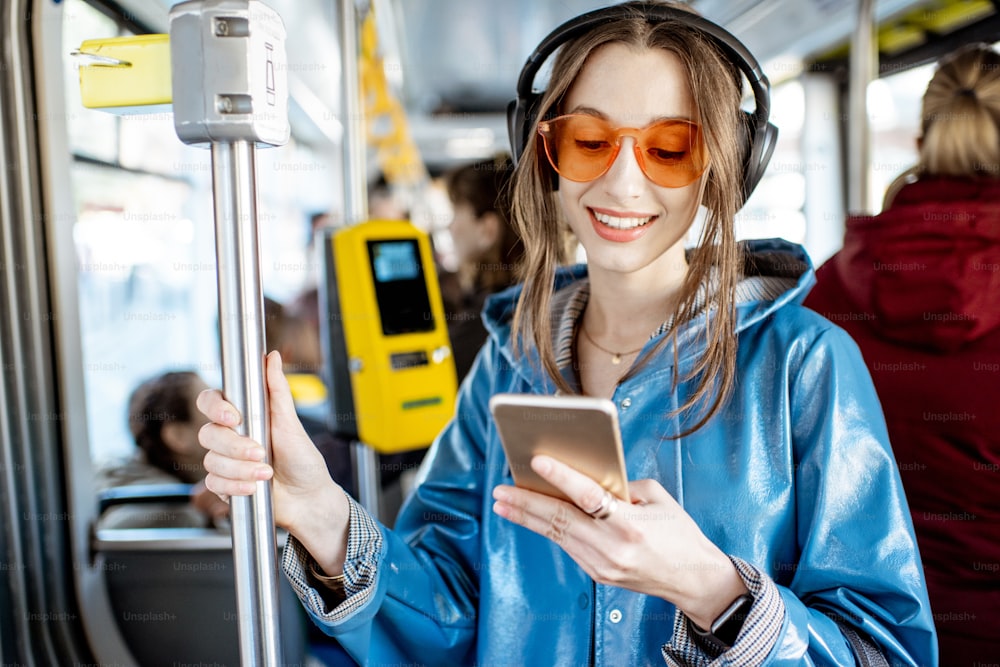 Junge stilvolle Frau, die öffentliche Verkehrsmittel benutzt, mit Kopfhörern und Smartphone steht, während sie sich in der modernen Straßenbahn bewegt