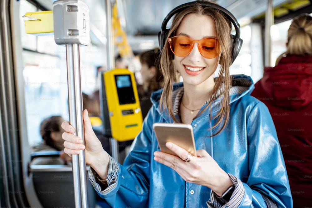 Mulher elegante nova usando o transporte público, de pé com fones de ouvido e smartphone enquanto se move no bonde moderno