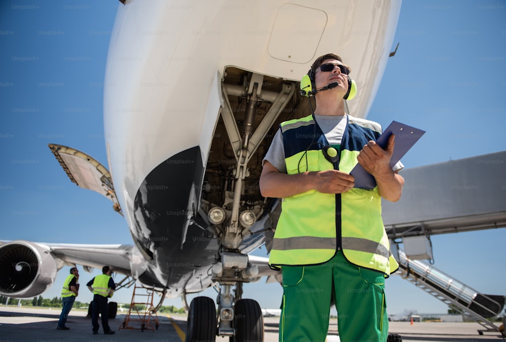 Preparación para el vuelo. Retrato de ángulo bajo de un hombre con gafas de sol sosteniendo documentos y posando cerca de un avión con la puerta abierta. Miembros de la tripulación de tierra parados detrás