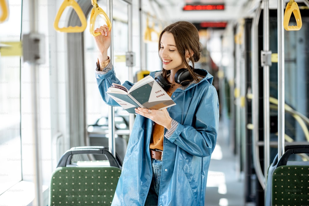 Junge Frau, die ein Buch liest, während sie in der modernen Straßenbahn steht, glücklicher Passagier, der sich mit bequemen öffentlichen Verkehrsmitteln bewegt