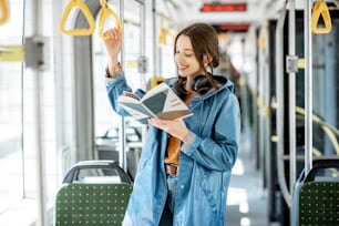 Mujer joven leyendo un libro mientras está de pie en el moderno tranvía, feliz pasajero que se mueve en cómodo transporte público