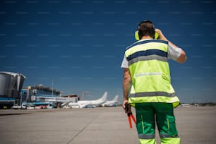 I hear you. Back view of man in signaling vest holding wands and checking the headset. Blue sky, runway, passenger planes and terminal on background