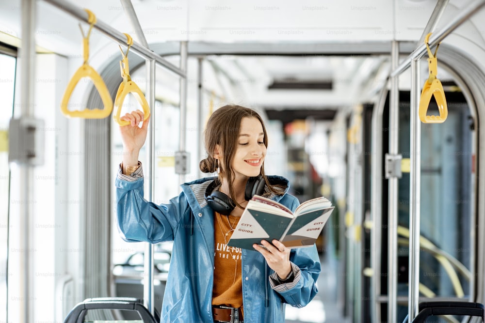 Junge Frau, die ein Buch liest, während sie in der modernen Straßenbahn steht, glücklicher Passagier, der sich mit bequemen öffentlichen Verkehrsmitteln bewegt