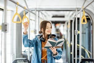 Young woman reading book while standing in the modern tram, happy passenger moving by comfortable public transport