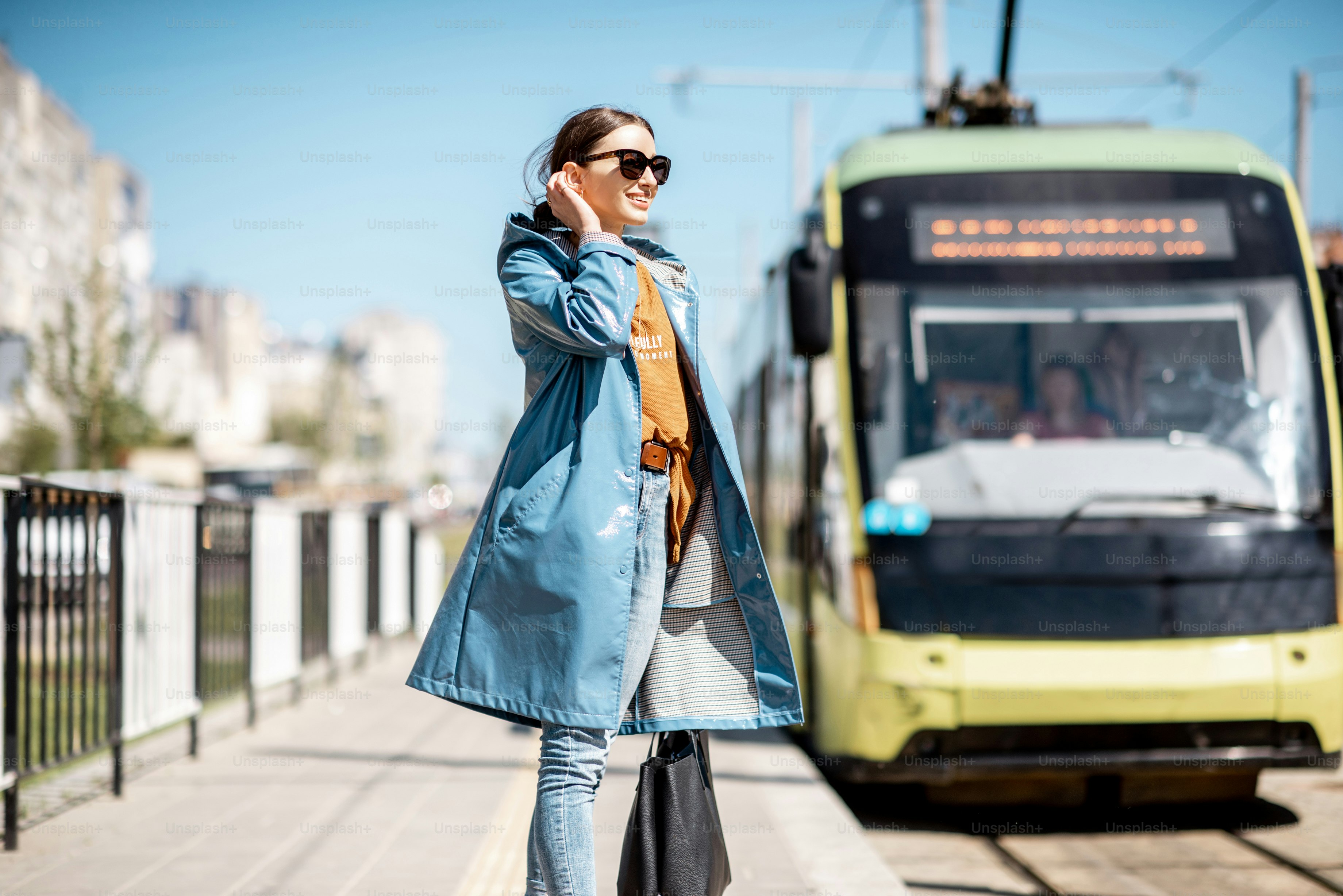 Junge Frau im blauen Mantel wartet im Freien auf der Station auf die Straßenbahn