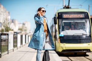Junge Frau im blauen Mantel wartet im Freien auf der Station auf die Straßenbahn