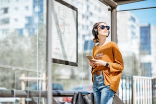 Young stylish woman waiting for the public transport while standing at the modern tram station outdoors