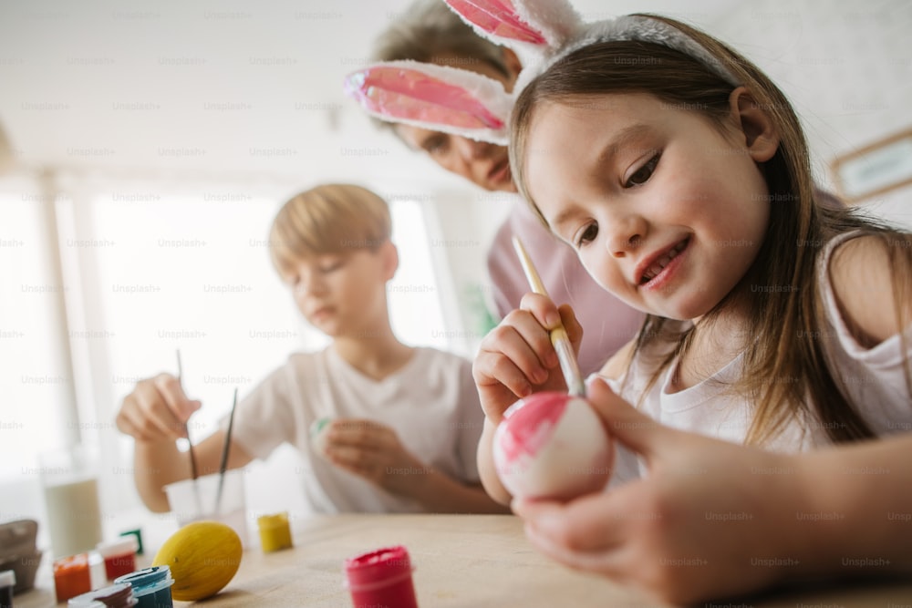 Cropped photo of smiling child with bunny ears on head coloring Easter egg indoors. She is sitting at table and holding paintbrush in arm. Family tradition concept