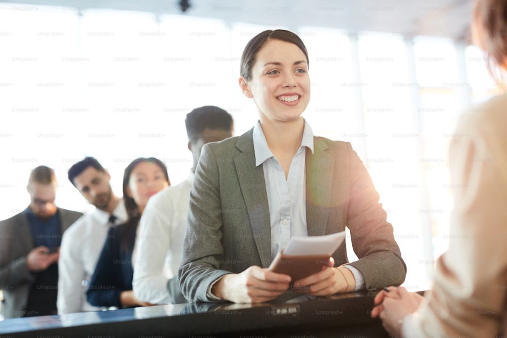 Successful young business professional in formalwear smiling at check-in manager by counter while consulting about flight details