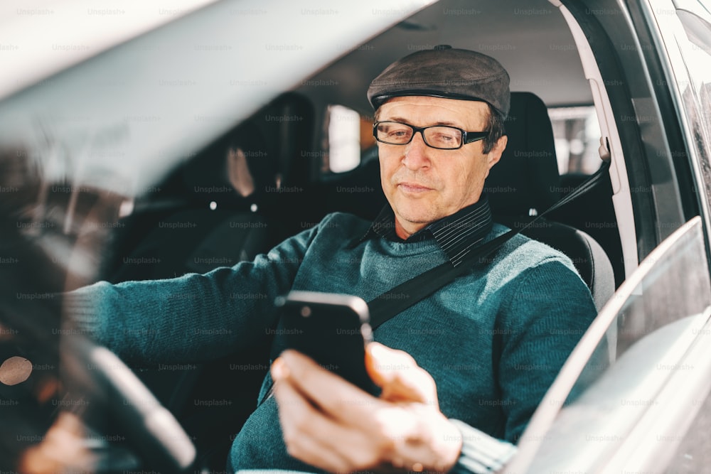 Hombre mayor caucásico serio con gorra en la cabeza y anteojos sentado en el automóvil y usando un teléfono inteligente. Ventana abierta.