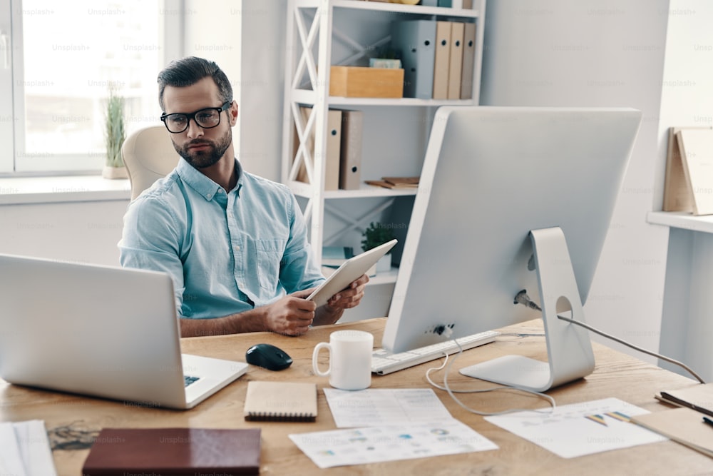 Junger moderner Geschäftsmann, der mit einem digitalen Tablet arbeitet, während er im Büro sitzt