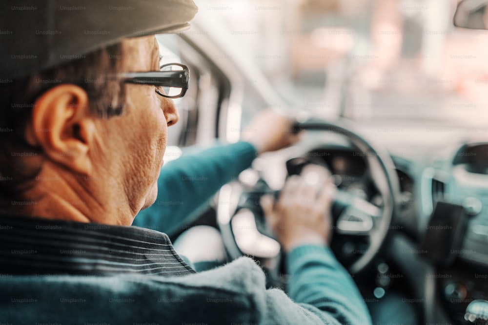 머리에 모자를 쓰고 안경을 쓴 백인 노인이 차를 운전하고 있다. 뒷좌석에서 찍은 사진.