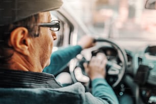 Uomo anziano caucasico con berretto in testa e occhiali da vista alla guida della sua auto. Foto scattata dal sedile posteriore.
