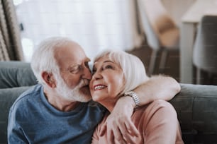 Love and tendance in any age. Close up portrait of happy smiling gray haired man kissing his wife with tender while enjoying time together on sofa