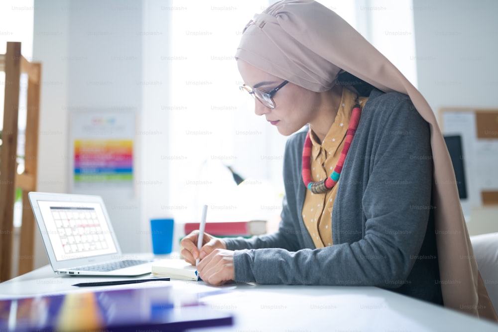 Teacher working. Muslim teacher wearing glasses making notes after work sitting near laptop