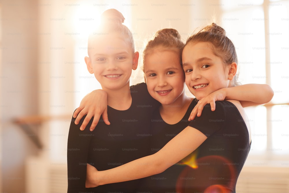 Portrait of three happy little girls embracing while posing in ballet studio lit by sunlight, copy space