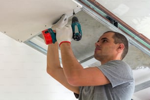 Construction worker assemble a suspended ceiling with drywall and fixing the drywall to the ceiling metal frame with screwdriver.
