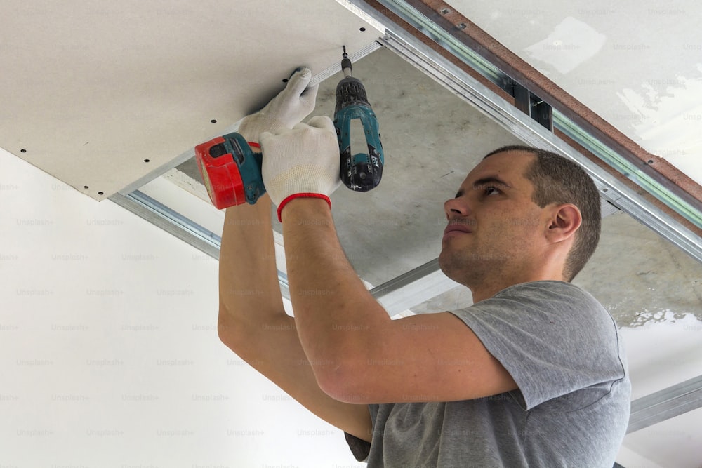 Construction worker assemble a suspended ceiling with drywall and fixing the drywall to the ceiling metal frame with screwdriver.