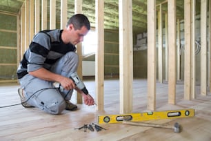 若いプロの労働者は、将来の壁のために木製フレームを取り付ける水平ドライバーとドライバーを使用します。再建中のオーク材の床を持つ屋根裏部屋の断熱部屋の内部。 改修と改善のコンセプト。