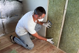 Worker in protective goggles and respirator insulating rock wool insulation in wooden frame for future house walls for cold barrier. Comfortable warm home, economy, construction and renovation concept