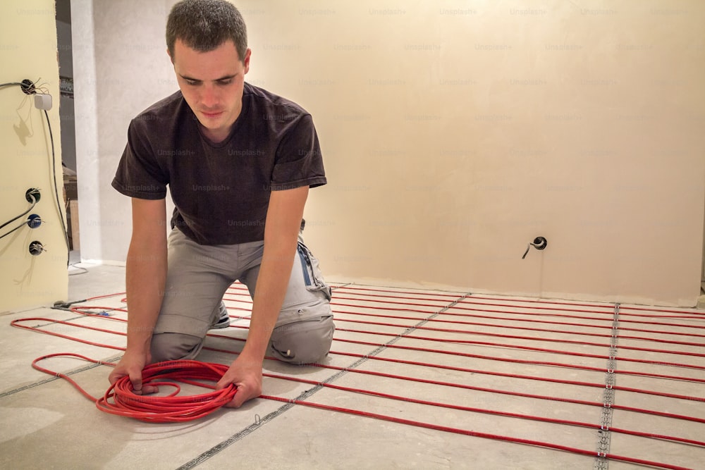 Electricista instalando cable eléctrico rojo calefactor en piso de cemento en habitación sin terminar. Renovación y construcción, concepto de hogar cálido y confortable.