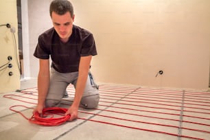 Électricien installant un fil de câble électrique rouge chauffant sur le sol en ciment dans une pièce non finie. Rénovation et construction, concept de maison chaleureuse et confortable.
