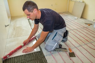 Young worker tiler installing ceramic tiles using lever on cement floor with heating red electrical cable wire system. Home improvement, renovation and construction, comfortable warm home concept.