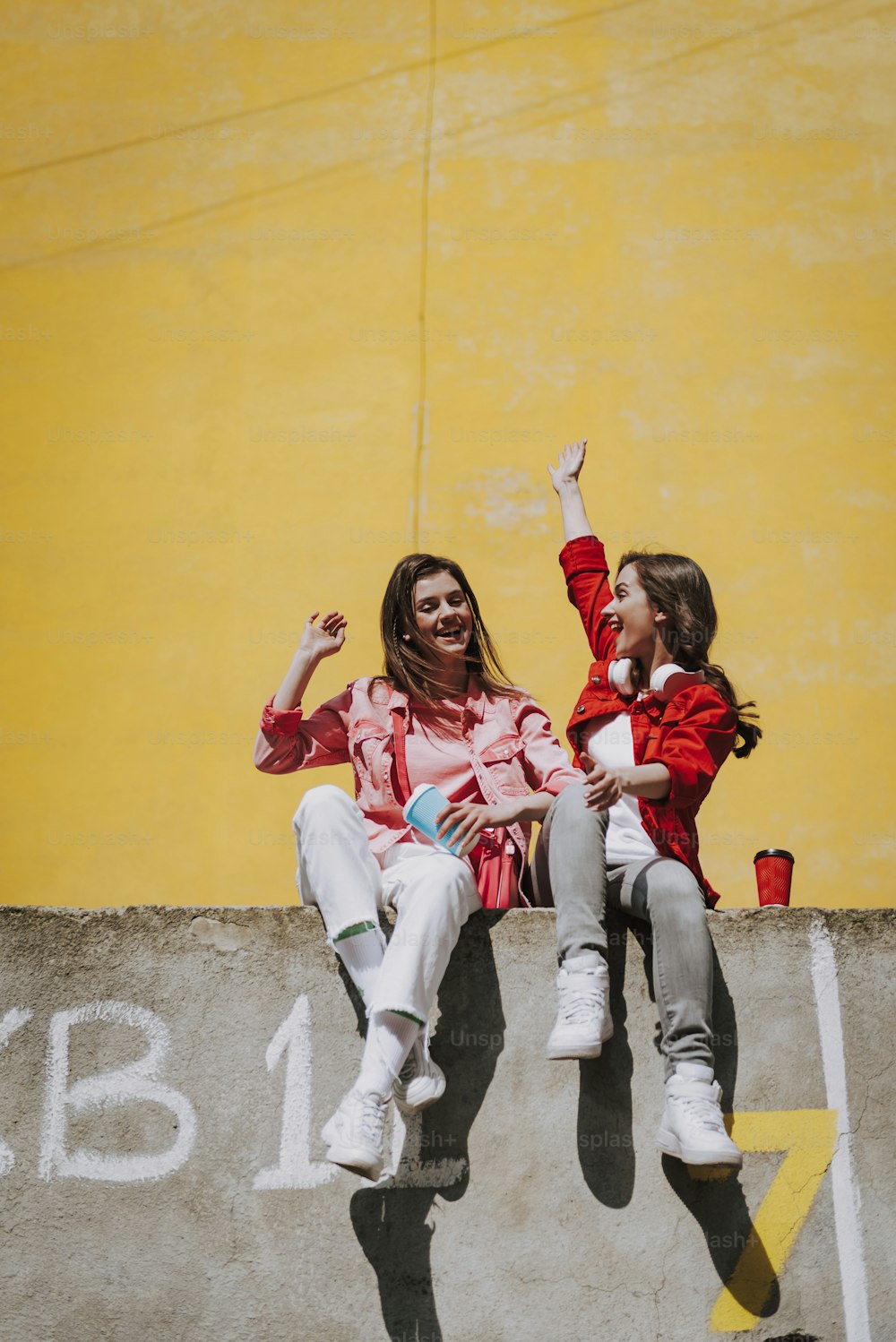 Urbanes Lifestyle-Konzept. Ganzkörperporträt zweier glücklicher junger Hipster-Damen, die sich gemeinsam auf der Brüstung mit einer Tasse Kaffee amüsieren