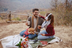 Couple enjoying picnic at autumn. Woman holding tablet, listening music over headphones and feeding her boyfriend while he holding cookies and mug. Dog lying down in man's lap.