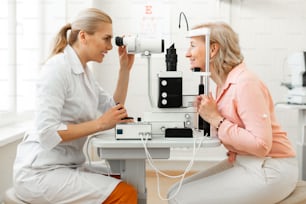 Using professional device. Smiling short-haired blonde woman in peachy shirt placing her head in metal frame during appointment