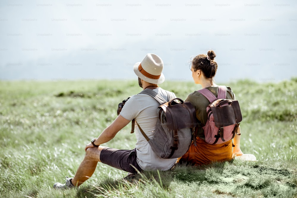 Young couple sitting on the green meadow, traveling with backpacks in the mountains during the summer time
