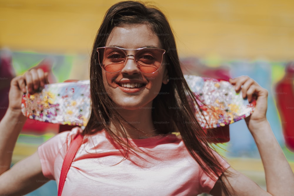 Estilo de vida urbano e atividade esportiva. Retrato de perto da jovem senhora hipster sorridente segurando penny board nas costas e ficando na vista da cidade