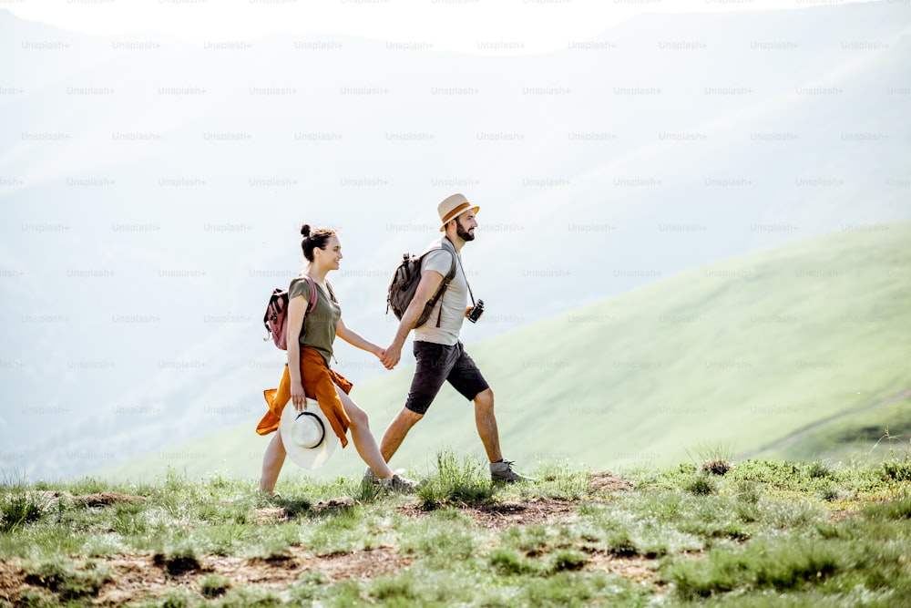 Beautiful couple walking with backpacks on the green meadow, while traveling in the mountains during the summer time