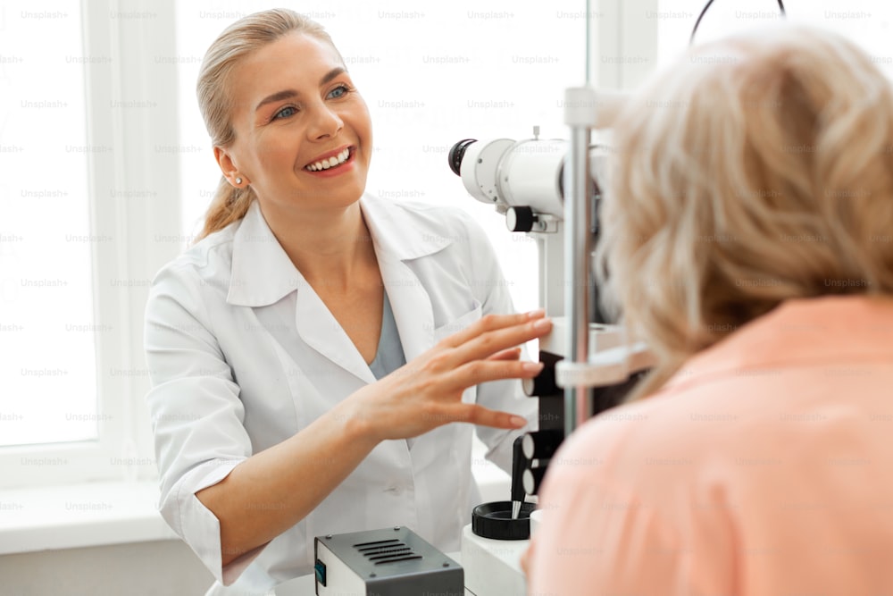 Contrôle de la vue. Ophtalmologiste positif riant en uniforme blanc travaillant avec un patient aux cheveux courts tout en utilisant le mécanisme
