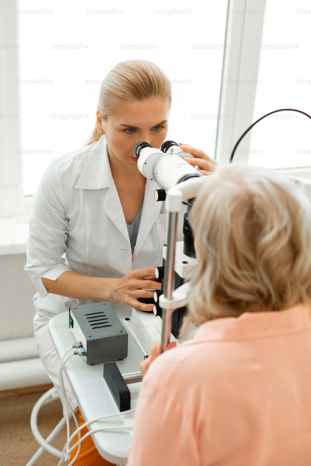 환자와 함께 일하기. 쌍안경을 들여다보며 금발을 묶은 세심한 의사가 환자의 눈 상태를 검사하고 있다