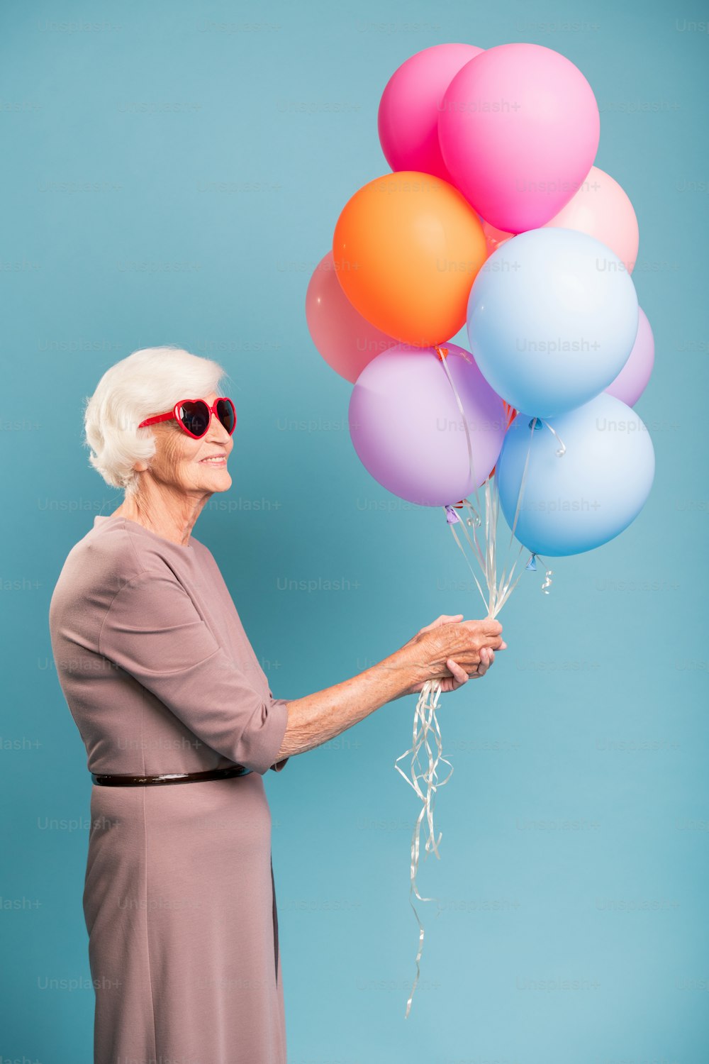 Elegant and gracious senior female holding bunch of colorful balloons while standing in studio