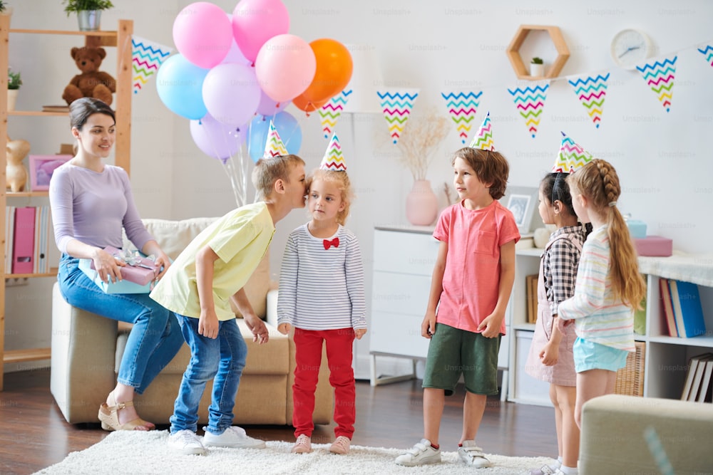 Group of little friendly kids playing whisper game in living-room while young woman with gifts sitting in armchair near by