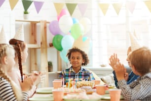 Portrait of smiling African-American boy wearing crown sitting at table while celebrating Birthday with friends, copy space