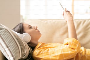 Profile view of young brunette woman in casualwear and headphones scrolling in smartphone while listening to music on couch