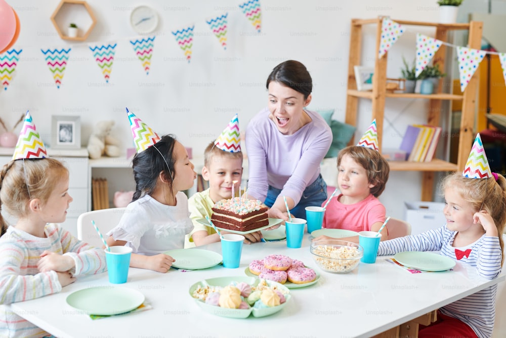 Mujer joven extasiada sosteniendo pastel de cumpleaños y mirando velas mientras una de las niñas pequeñas las sopla junto a la mesa servida entre amigos