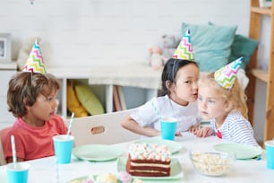 One of cute girls in birthday caps whispering something to her friend while sitting by festive table with boy near by
