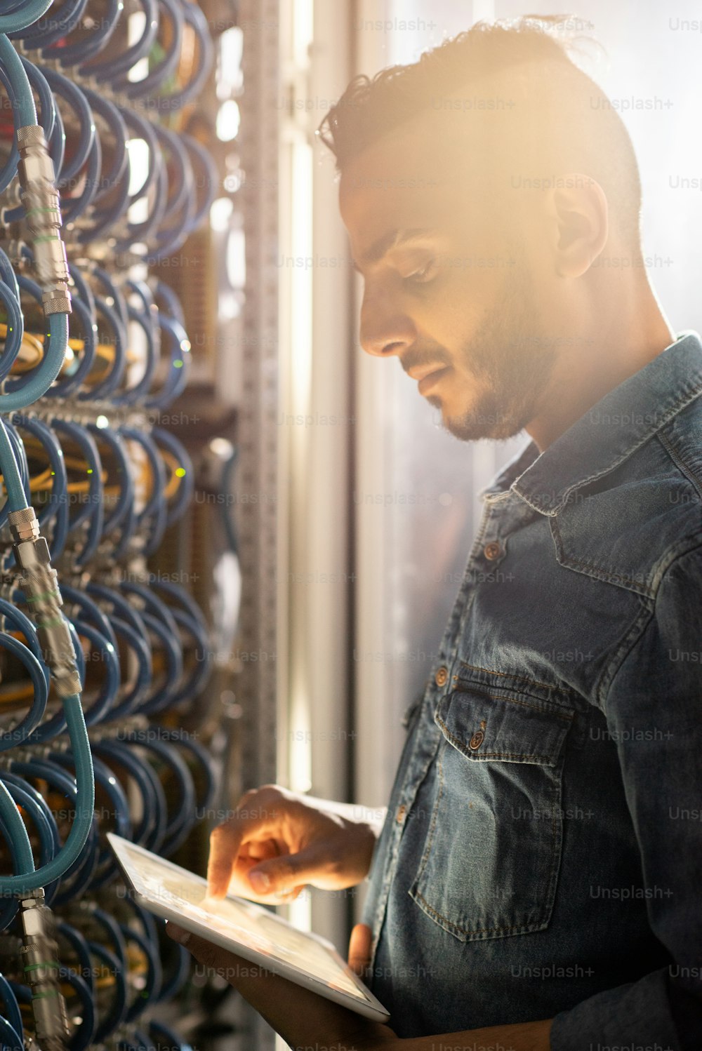 Serious thoughtful young middle-eastern IT engineer in denim jacket standing by cabinet of database server and viewing test results on tablet