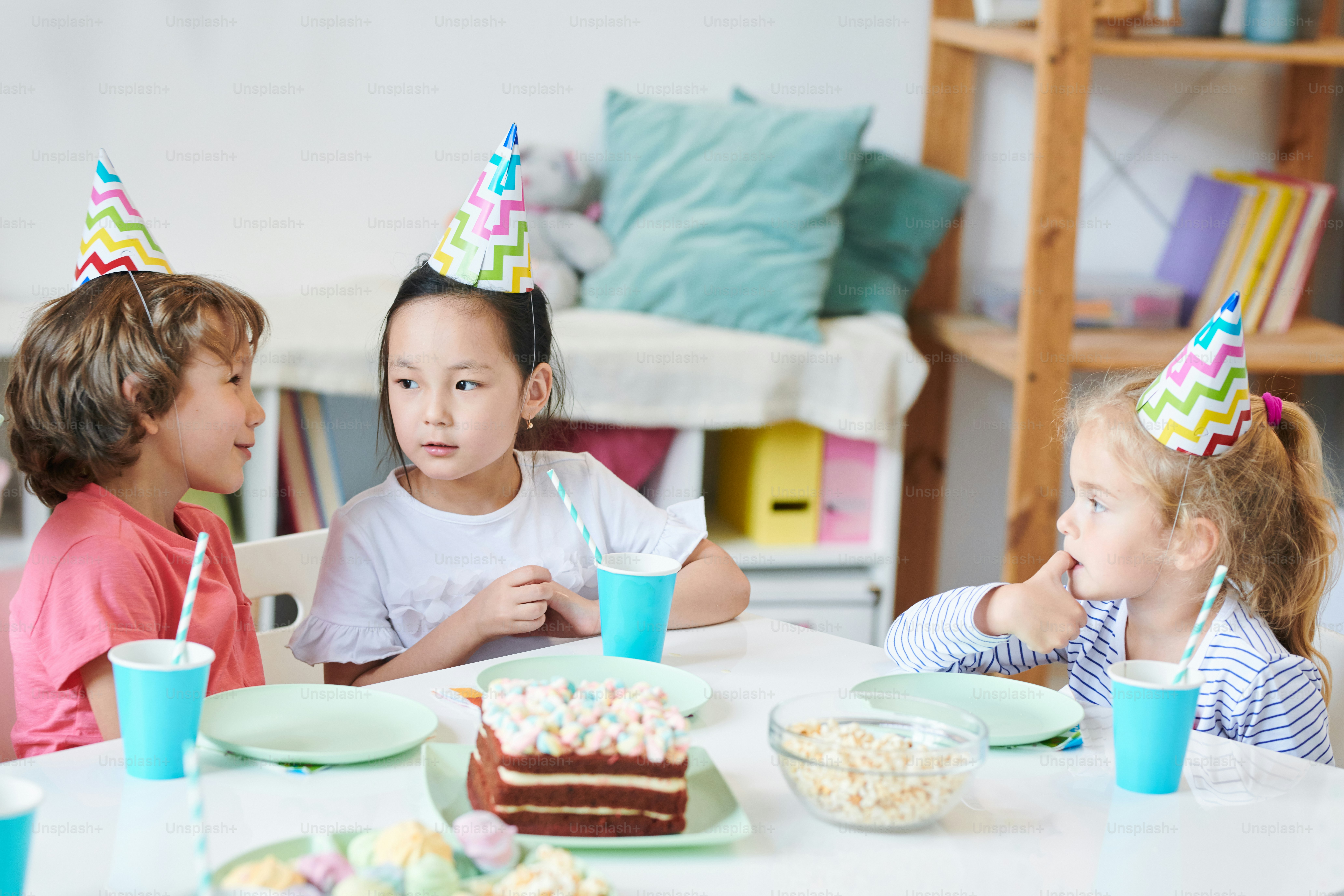 Lindo niño diciéndole algo a una linda niña asiática en la gorra de cumpleaños junto a la mesa festiva mientras toma bebidas y postre