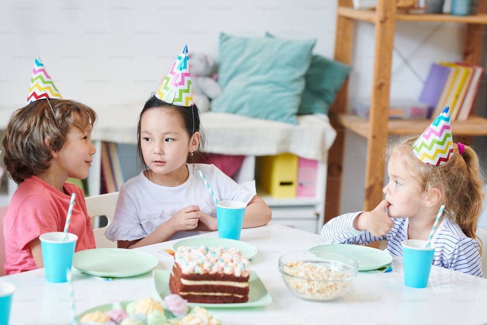 Netter kleiner Junge, der einem hübschen asiatischen Mädchen mit Geburtstagsmütze am festlichen Tisch etwas erzählt, während er Getränke und Dessert trinkt