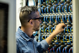 Joven ingeniero de mainframe árabe serio en gafas de pie junto al gabinete del servidor y analizando el sistema de almacenamiento de bases de datos