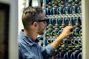 Jeune ingénieur mainframe arabe sérieux en lunettes debout près de l’armoire de serveur et analysant le système de stockage de base de données